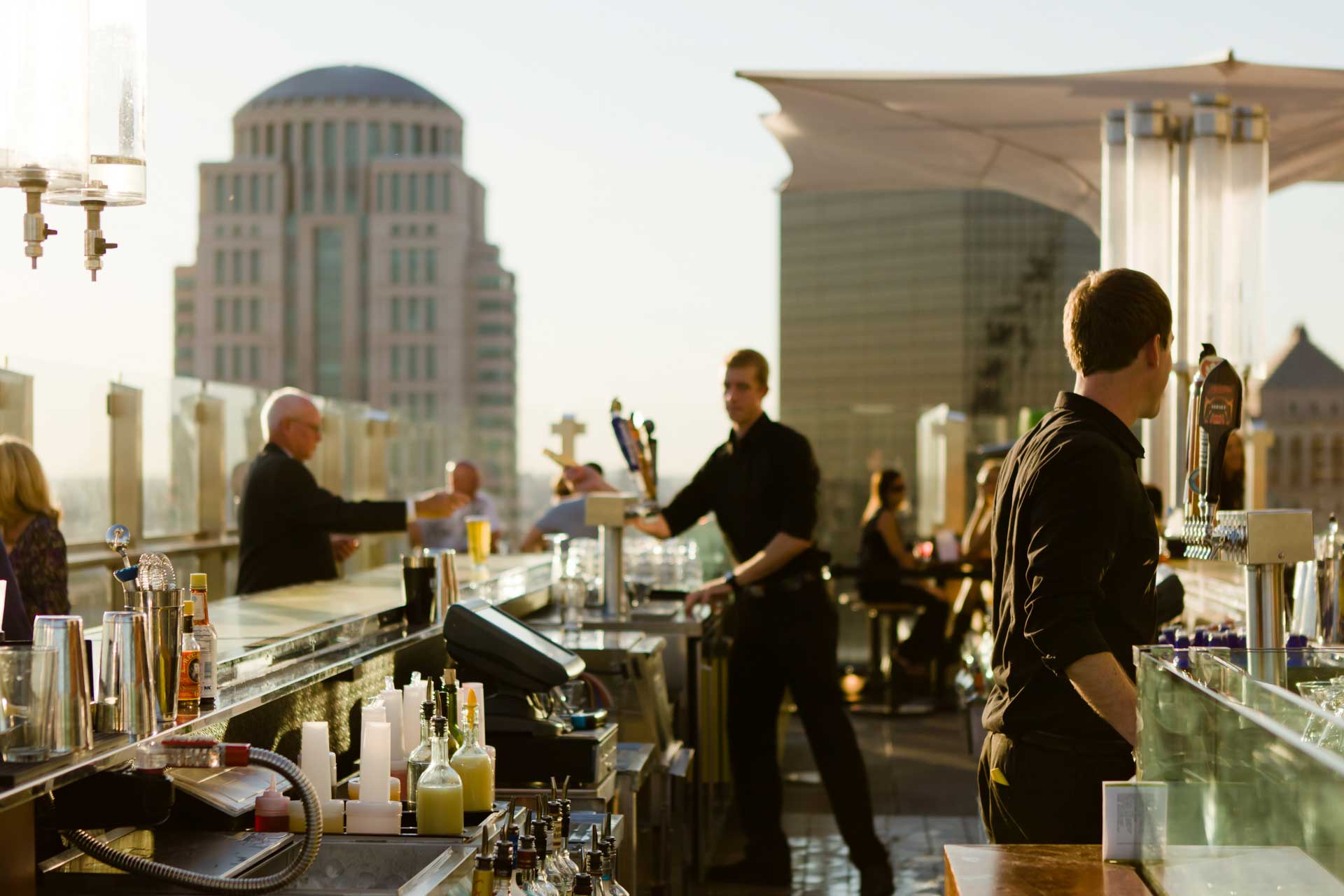 A group of people socializing at a rooftop bar, enjoying drinks and the city skyline in the background.