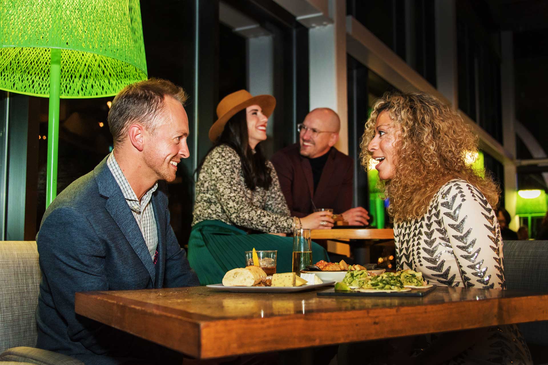 Four people seated at a table, illuminated by a green lamp, engaged in conversation.