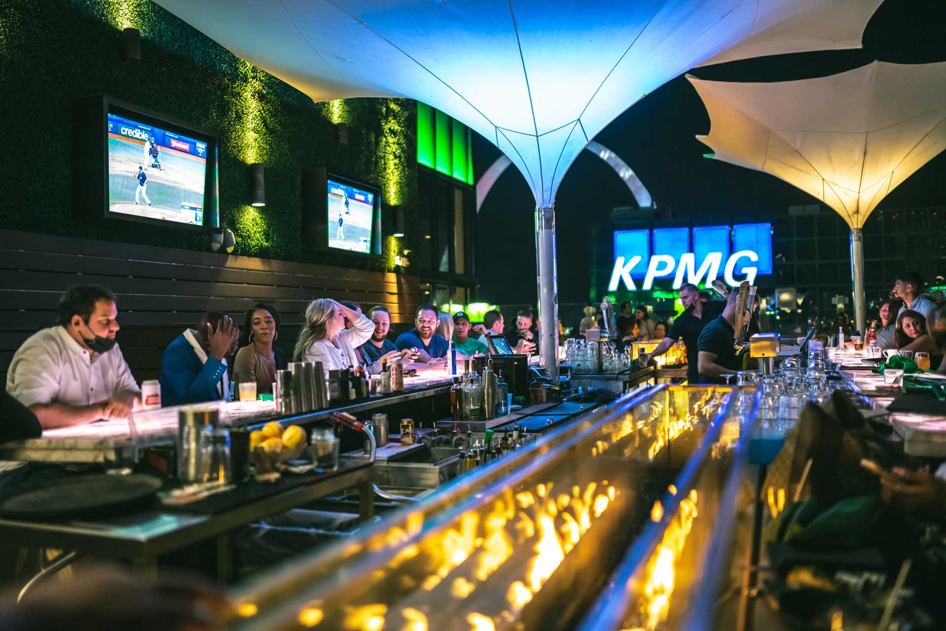 A group of people socializing at a bar, with a television screen displaying a sports game in the background.